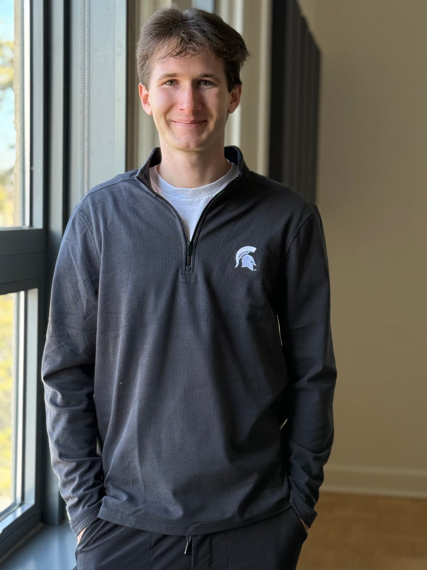 Man standing indoors, wearing a dark gray quarter zip with a white Spartan Helmet logo embroidered on the upper left chest.