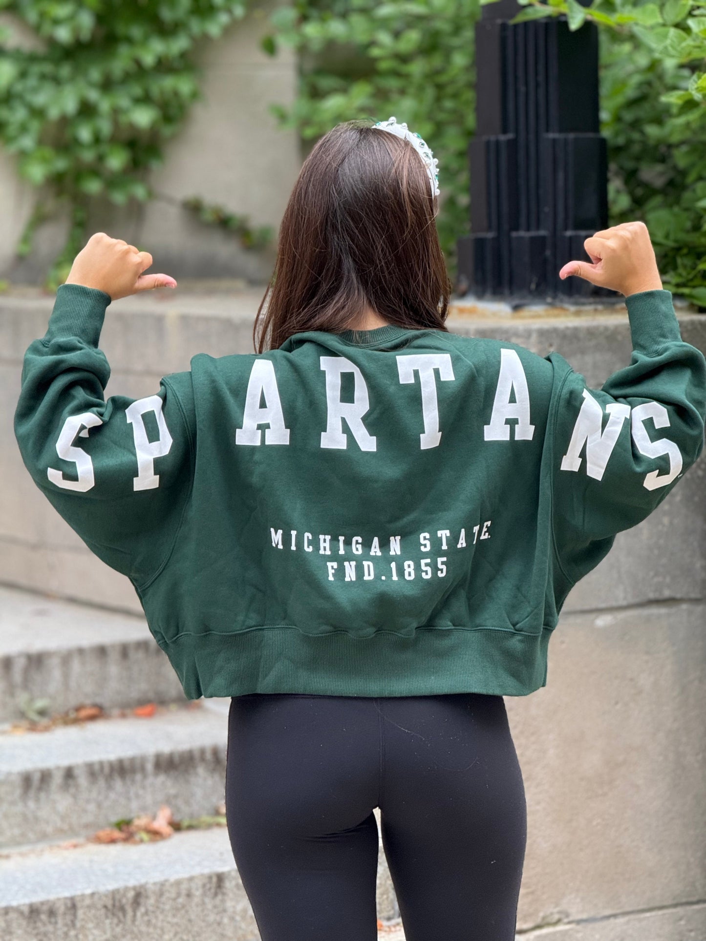 Woman standing outdoors, showing off the back of a green cropped crewneck sweatshirt with a "Spartans" wordmark across the arms and upper back and a "Michigan State FND. 1855" text below it, both printed on in white.