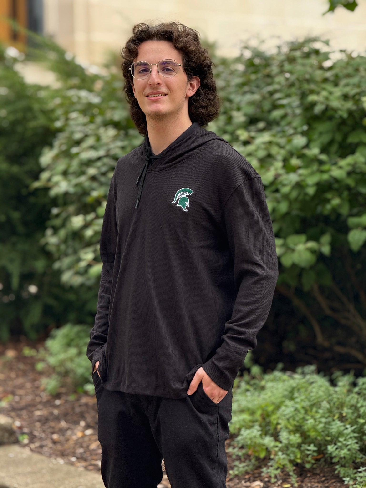 Person standing outdoors with hands in pockets, wearing a black hoodie with a Michigan State Green Helmet logo on the left chest. Background includes green bushes and plants. Face is blurred for privacy.