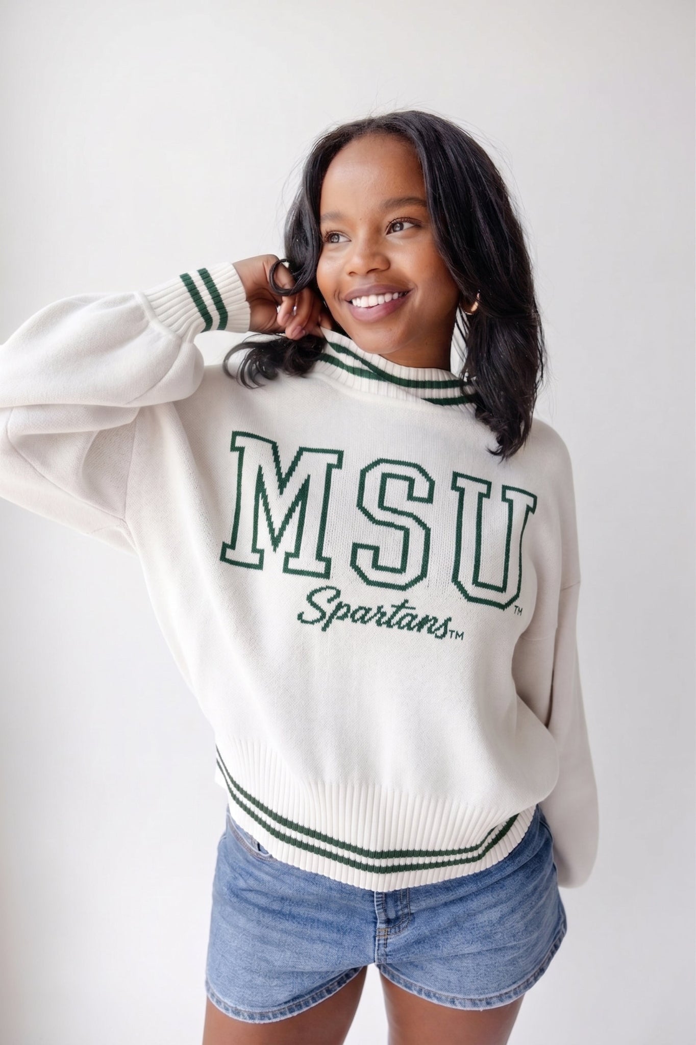 Woman wearing a white Michigan State University sweater with green “MSU Spartans” lettering and striped ribbed trim, paired with denim shorts.