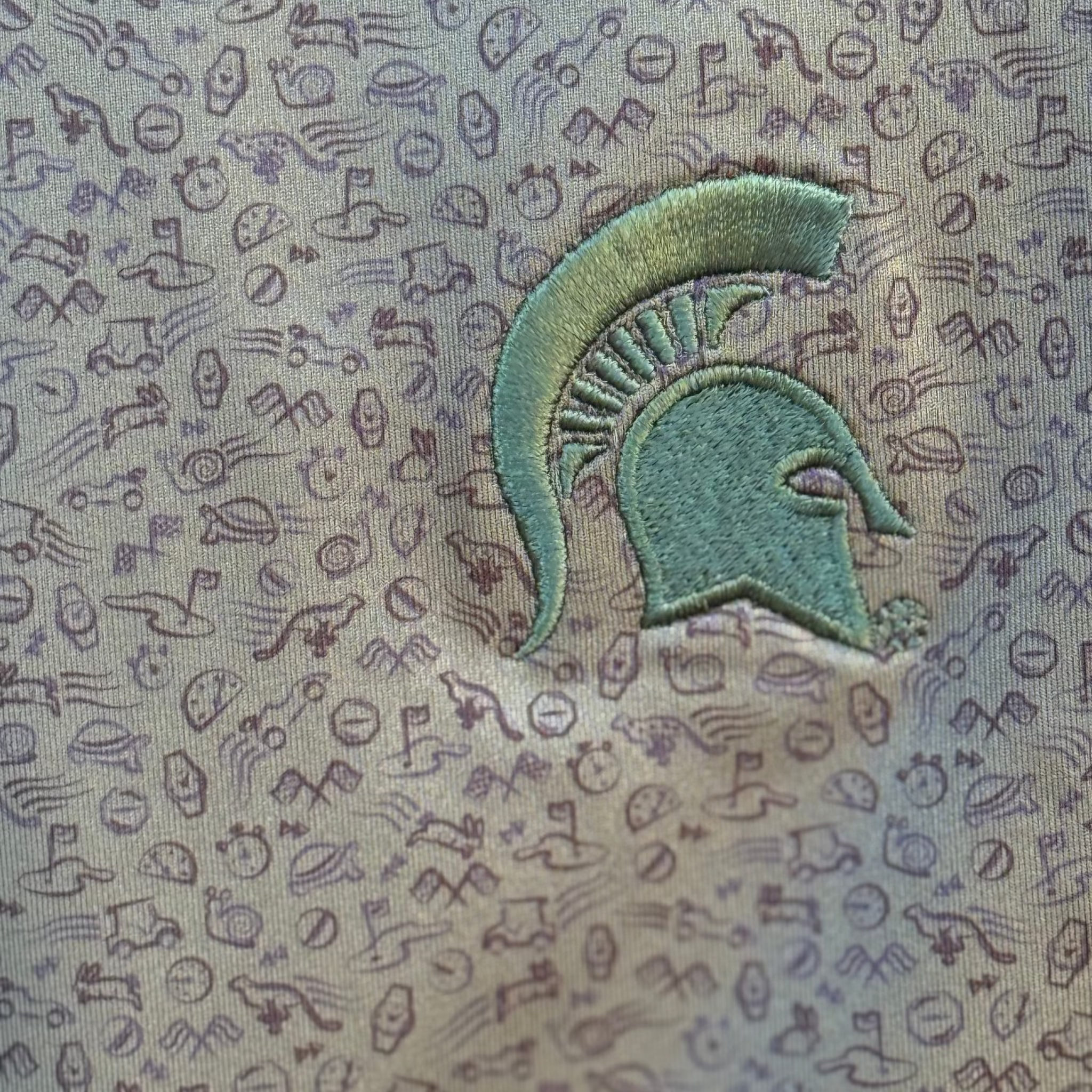 Close up on the dark green polo, showing the green Spartan helmet logo on a golf themed patterned background.