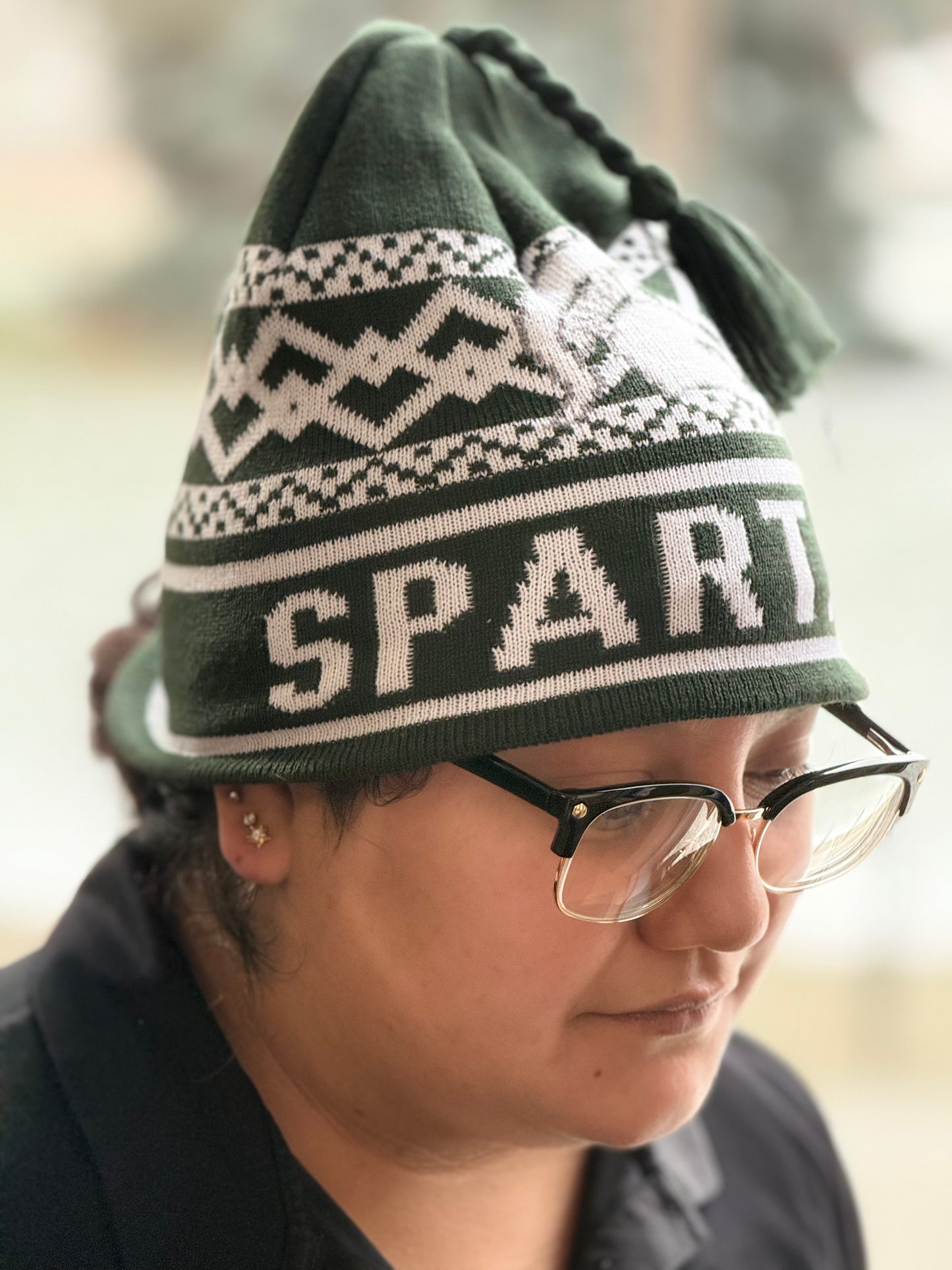 Side view of a green and white knit beanie with tassel, featuring a winter-inspired pattern and bold “Spartans” lettering across the cuff. Modeled on a person standing outdoors.