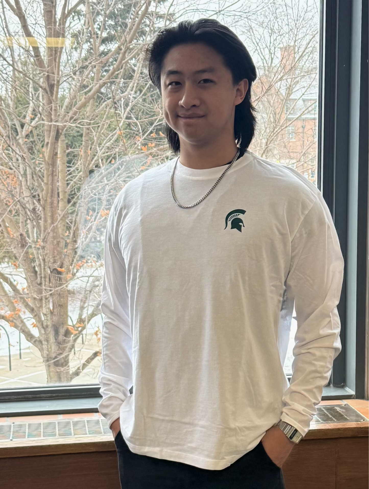 Person wearing a white long-sleeve shirt with a green screen printed Spartan helmet logo on left chest, standing in front of a window with bare trees outside.