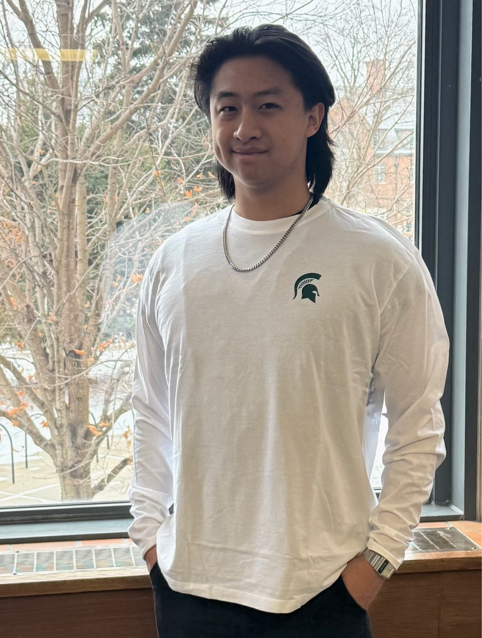 Person wearing a white long-sleeve shirt with a green screen printed Spartan Helmet logo on left chest, standing in front of a window with bare trees outside.