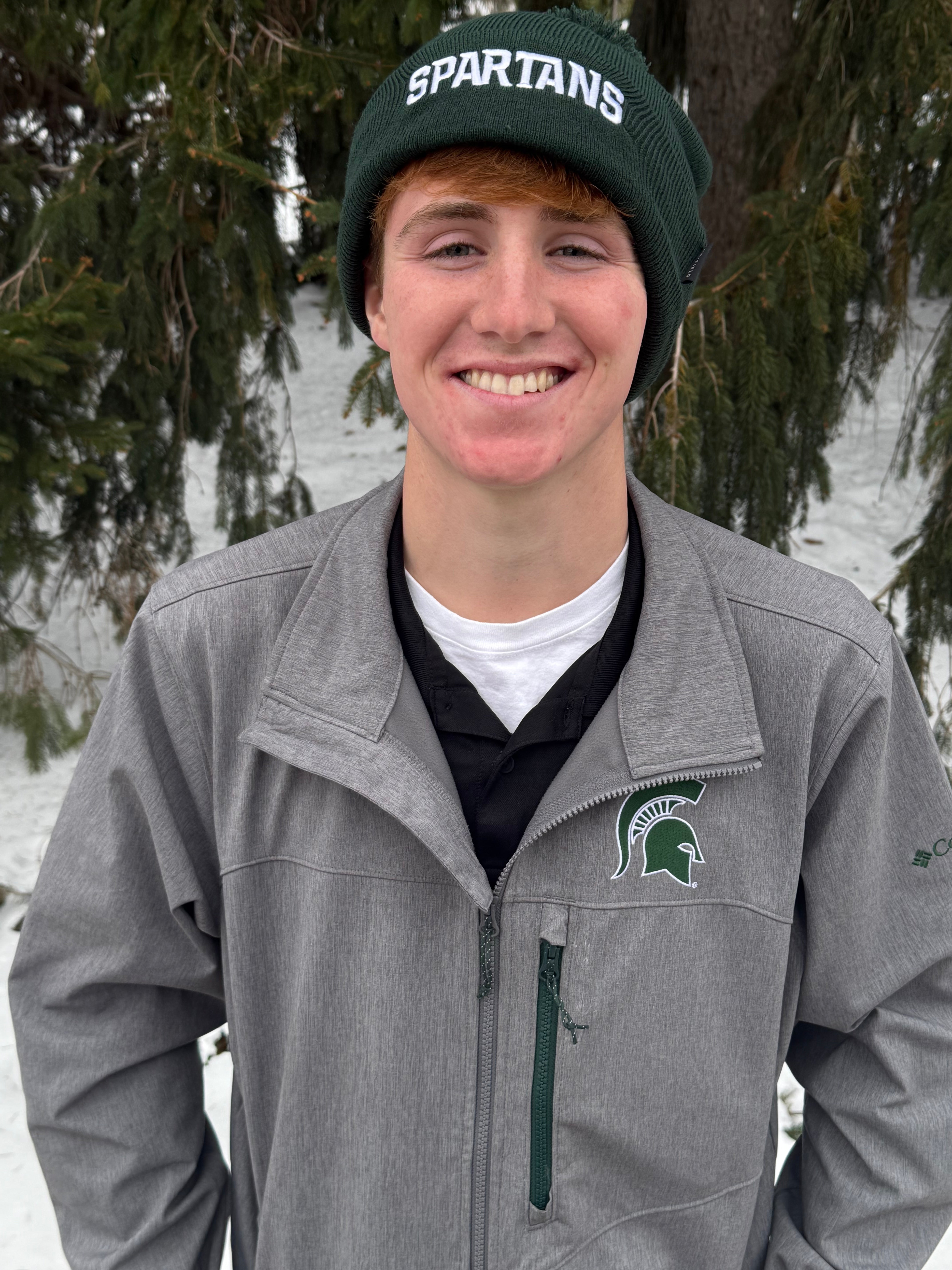 Man standing outdoors, wearing a green cuffed beanie with a green pom at the top and a white Spartans wordmark embroidered on the cuff.