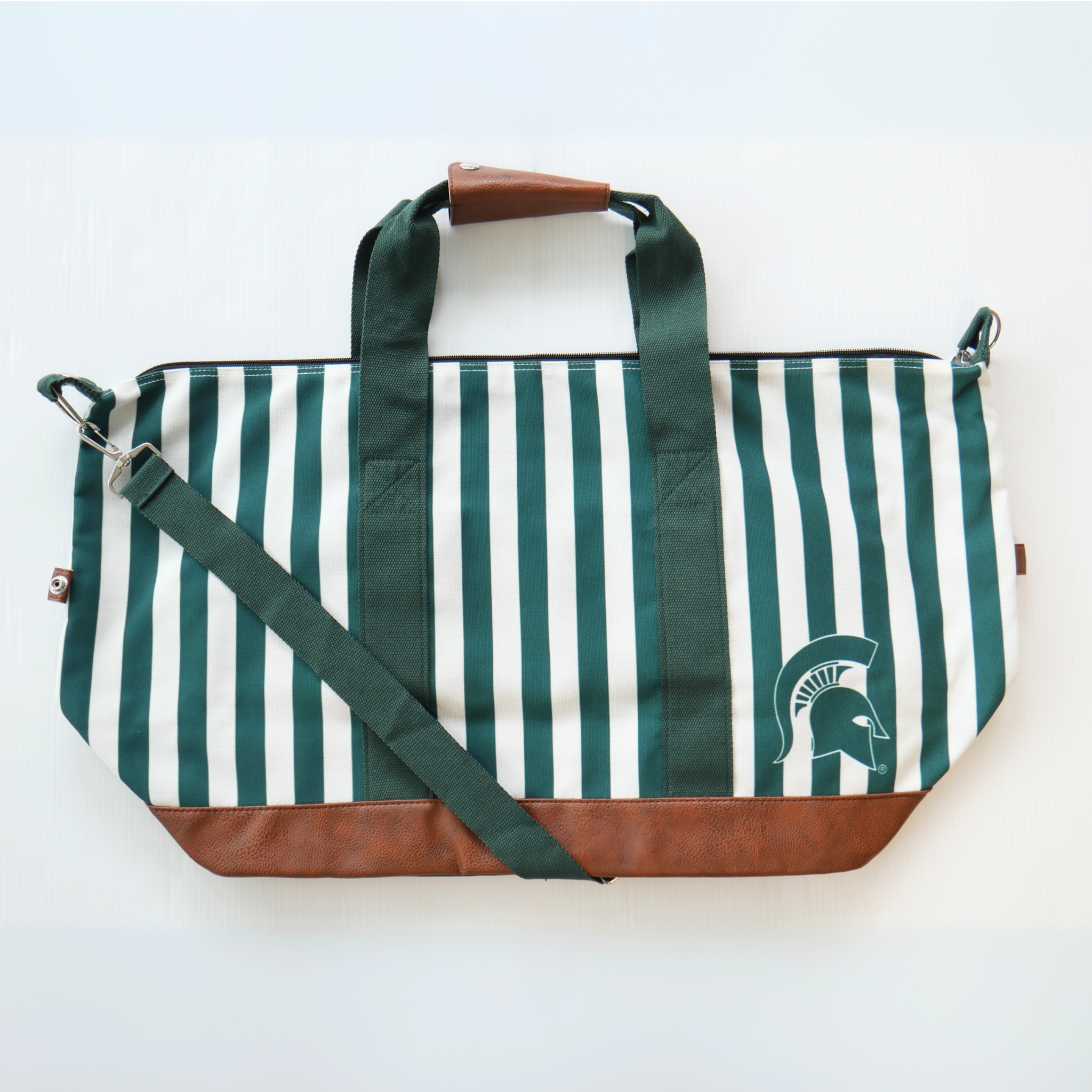 A green and white vertical striped canvas tote bag with a faux brown leather bottom and strap handles. The bag also has a green crossbody strap. In the lower left corner of the tote bag is a green MSU spartan helmet logo.