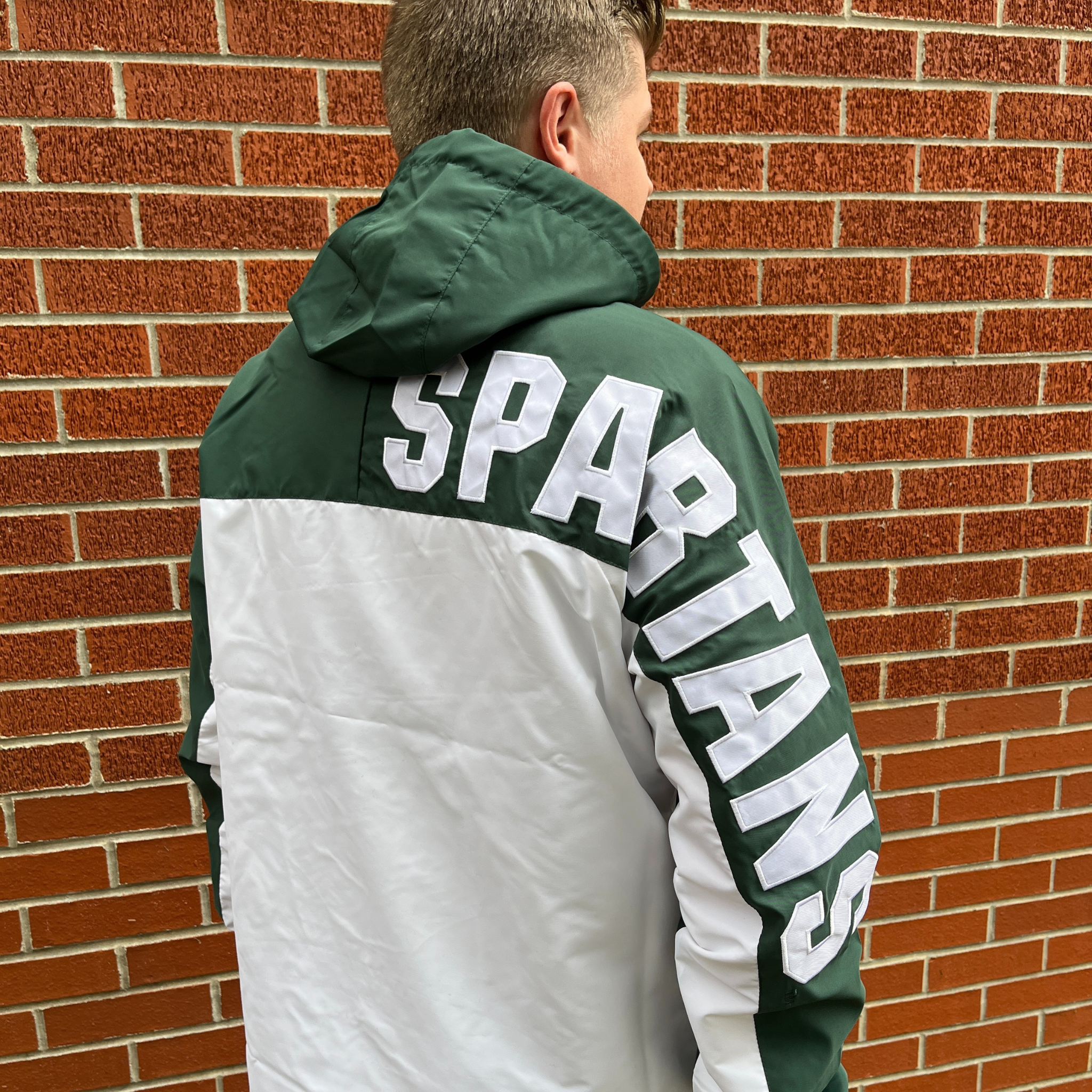 A person wearing a green and white hooded rain jacket. The jacket has the MSU spartan helmet logo in white on the left chest, with Spartans in all caps down the side of the right sleeve.