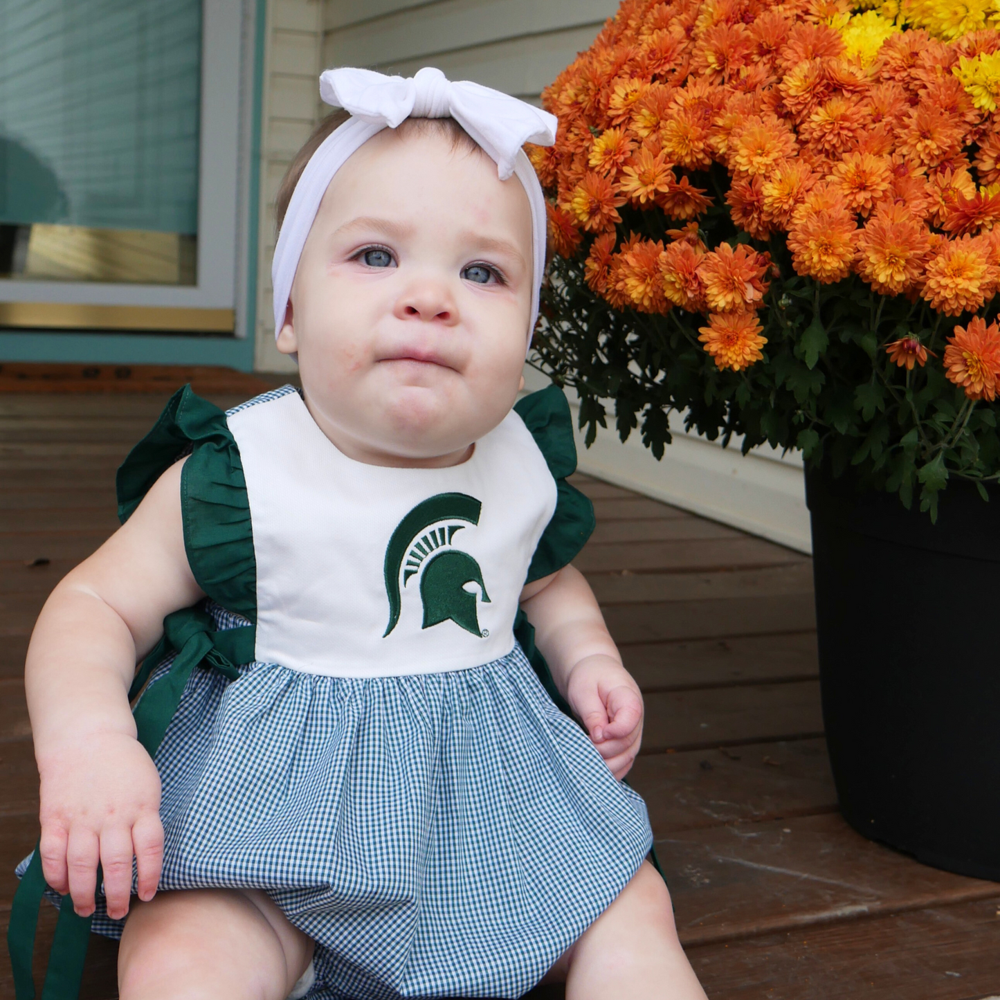 A baby wearing a sleeveless baby jumper next to a large bouquet of orange flowers. The green and white plaid bottom has a bubble effect in shape. The top is white with dark green ruffle trim ending in bows at the hip. A dark green Spartan Helmet is printed on the center chest.