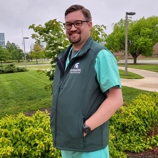 Person showing the left side of a green zippered vest with a white embroidered College of Osteopathic Medicine logo on the left upper chest.