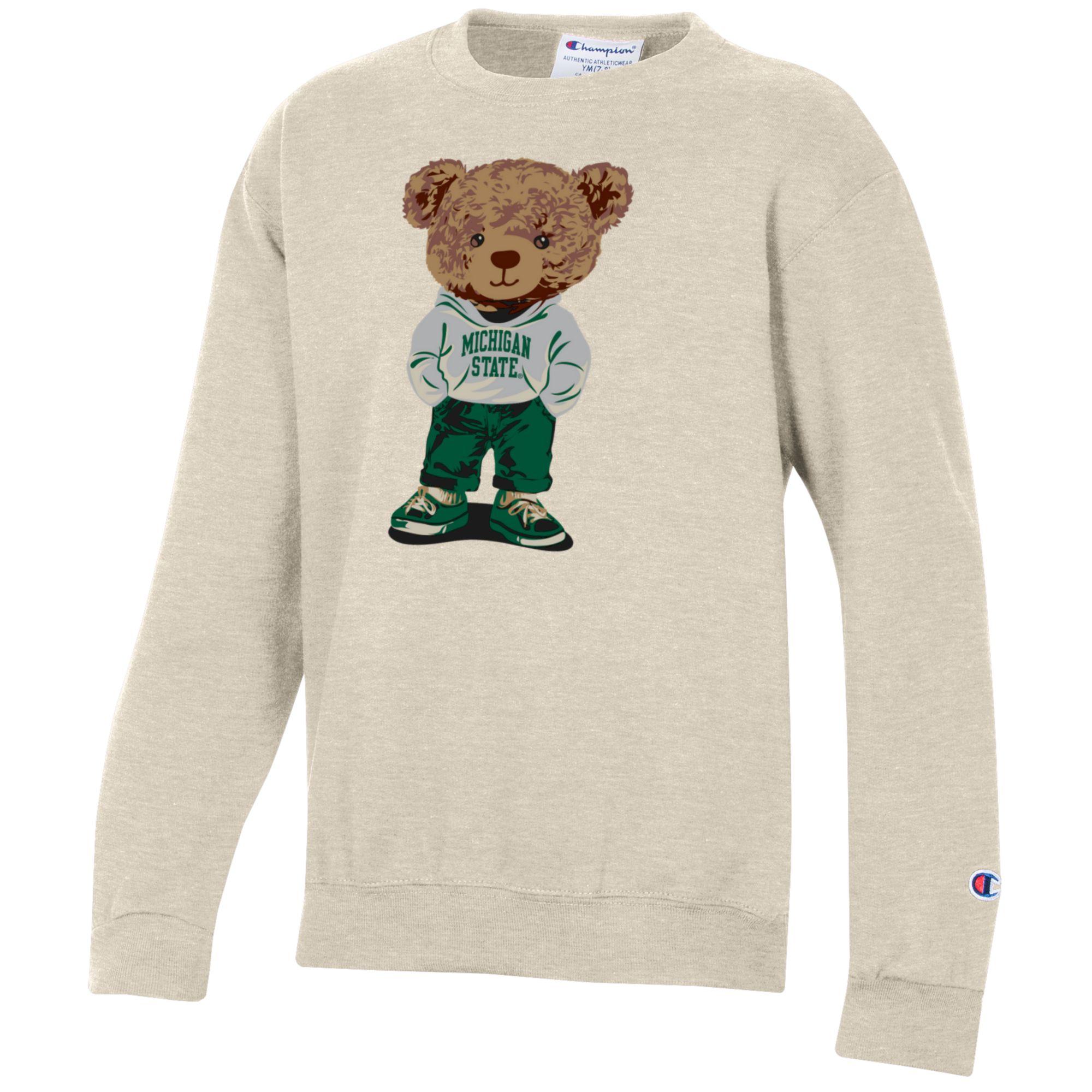Light beige crewneck sweatshirt with a teddy bear graphic that is wearing a sweatshirt with "Michigan State" written on it, green pants, and green and white shoes. The classic Champion logo is embroidered on the lower left cuff.