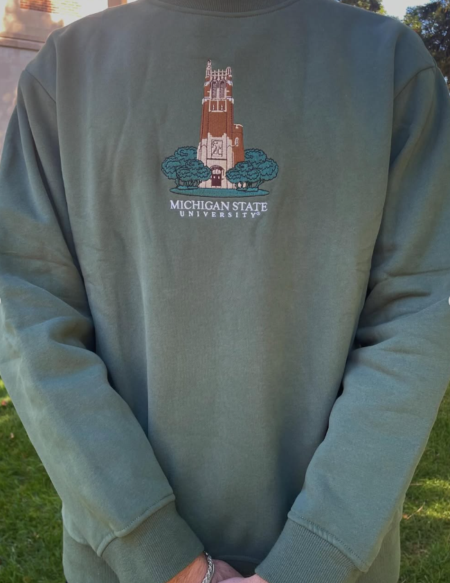 Person standing outdoors, wearing a green crewneck sweatshirt with the Michigan State University Beaumont Tower logo embroidered on the front and a 'Michigan State University' wordmark below it in white.