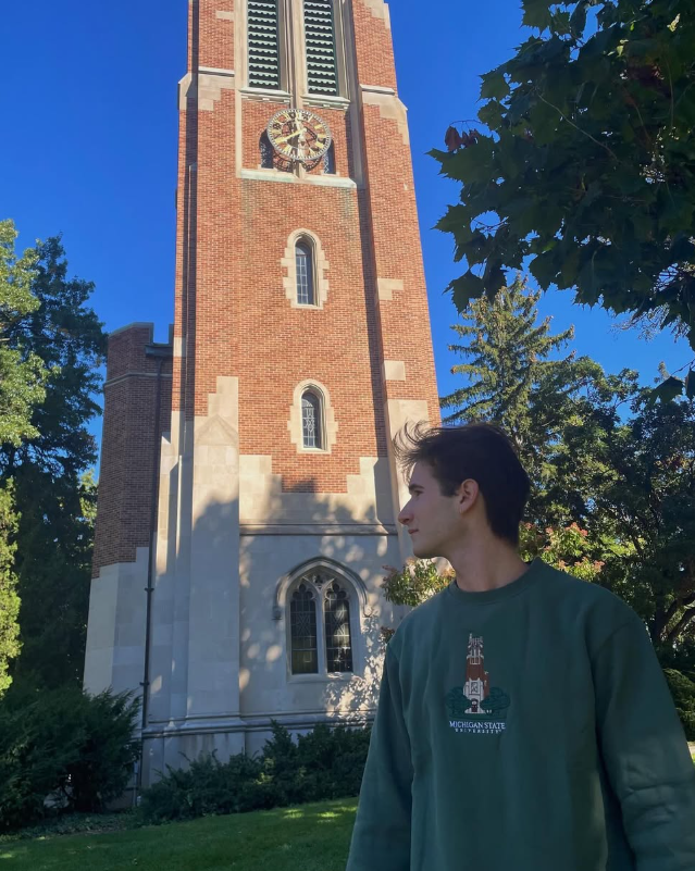 Person standing in front of the Michigan State Beaumont tower, wearing a green crewneck sweatshirt with the Michigan State University Beaumont Tower logo embroidered on the front and a 'Michigan State University' wordmark below it in white.