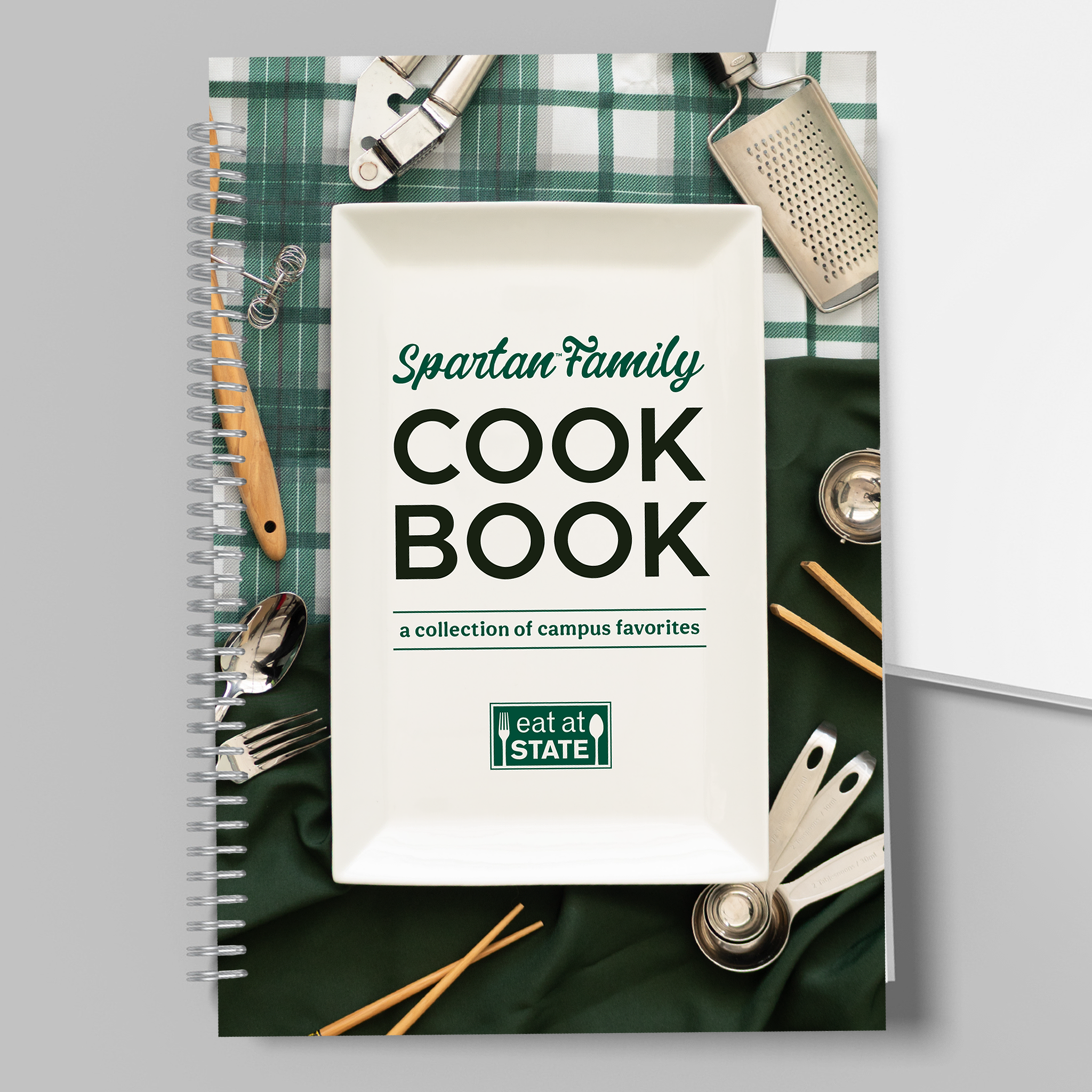 Cover of the The Spartan Family Cookbook. The text on the cover is shown on a plate on top of green and white tablecloths with kitchen utensils placed around.