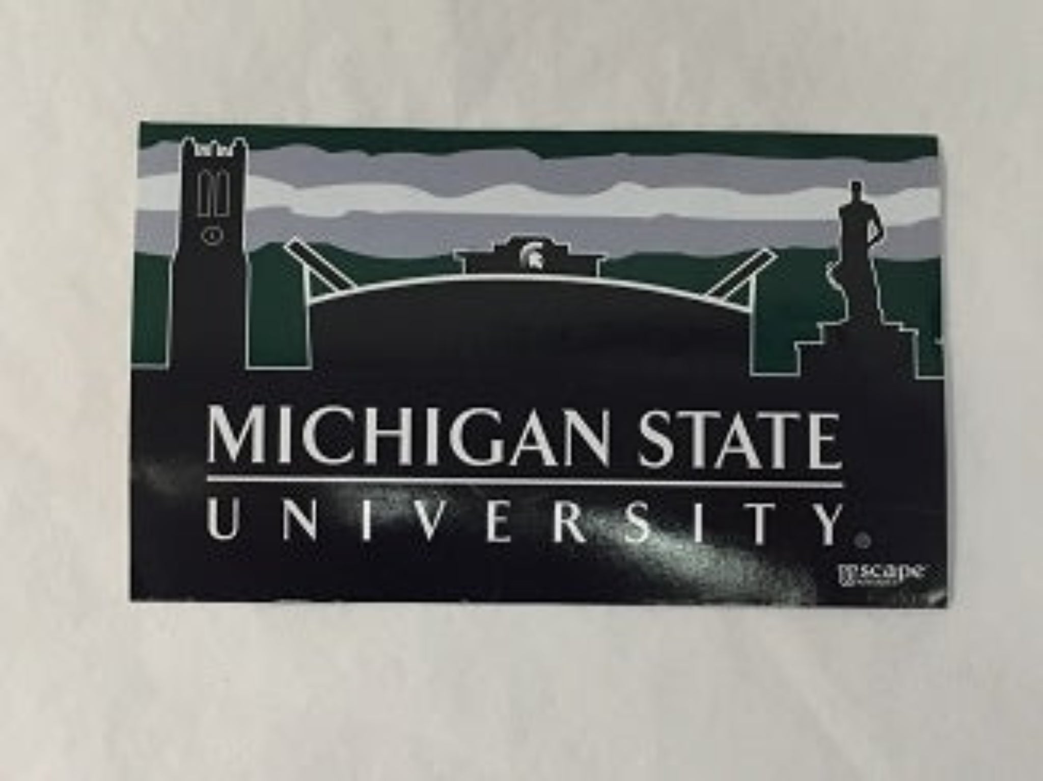 Sticker featuring 'MICHIGAN STATE UNIVERSITY' in white capital letters on a black background. Above the text are the illustrated landmarks: Beaumont Tower on the left, Spartan Stadium with the Spartan helmet logo in the center, and The Spartan statue on the right. The background includes stylized green and gray stripes.