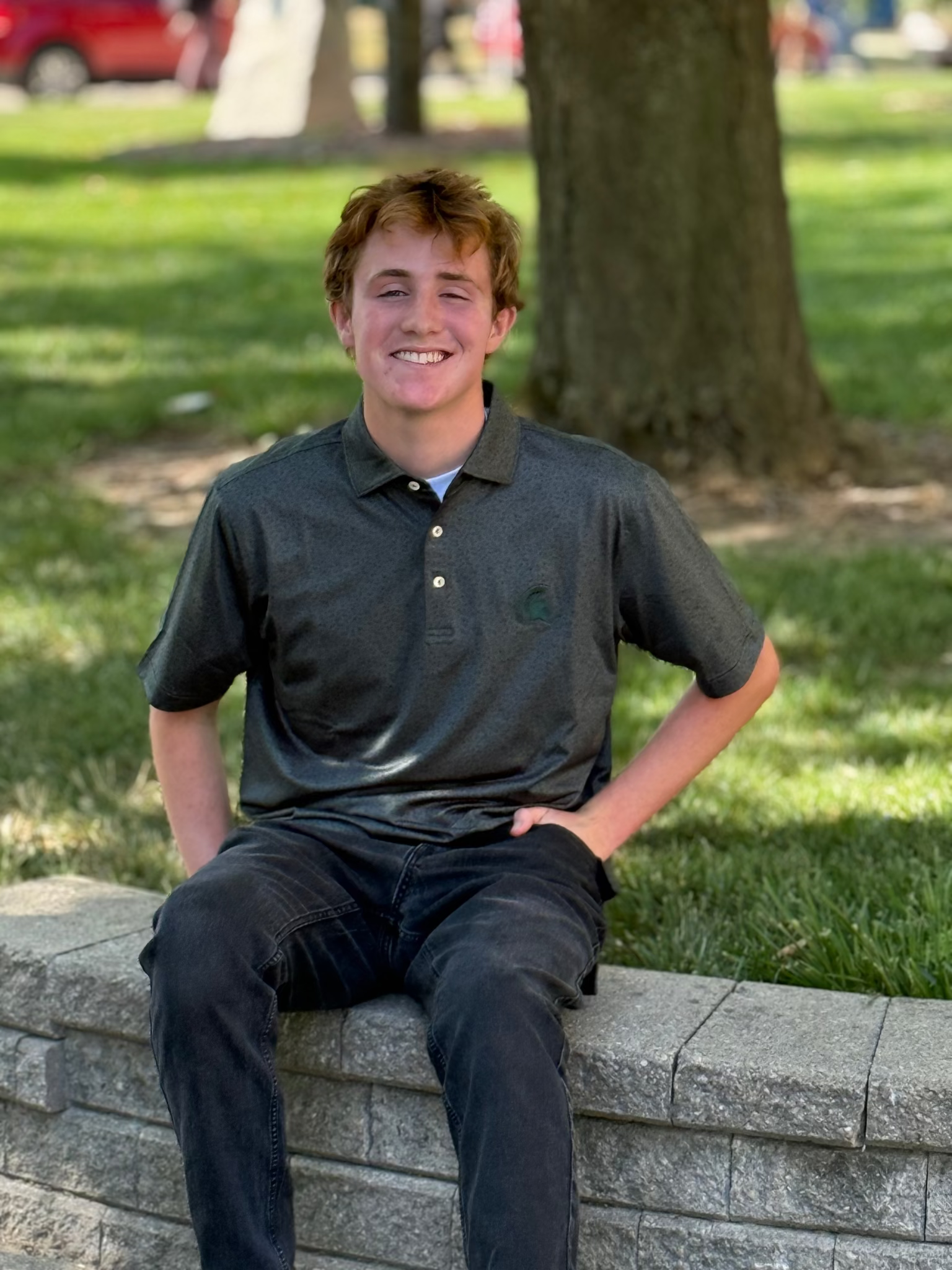 Person sitting on a stone wall, wearing dark green polo with small golf icons and symbols printed all over and a green Spartan helmet logo on the left upper chest.