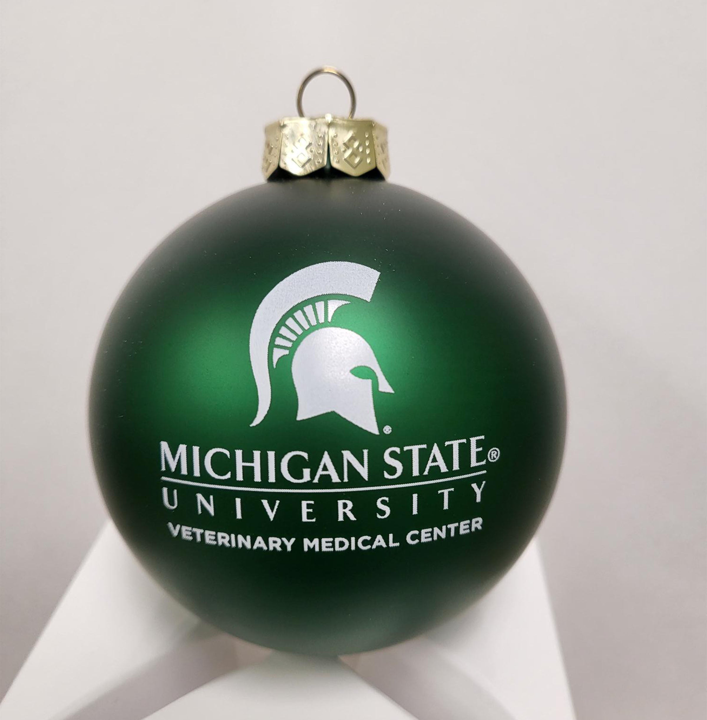 Green ornament with a gold tone top, white Spartan helmet logo and "Michigan State University Veterinary Medical Center" printed in white in the center.