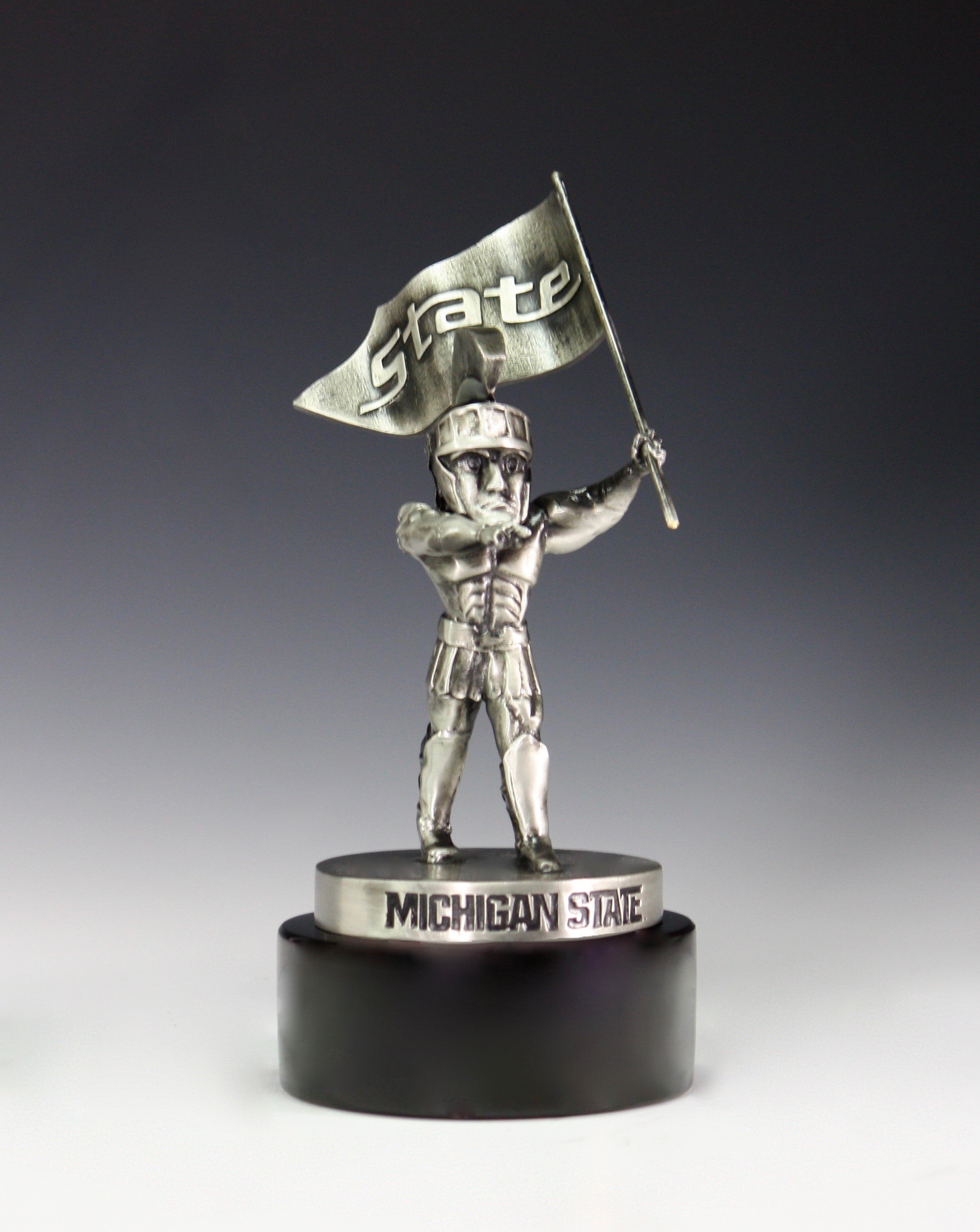 A silver Sparty statue holding a flag that reads "State" on a black base.