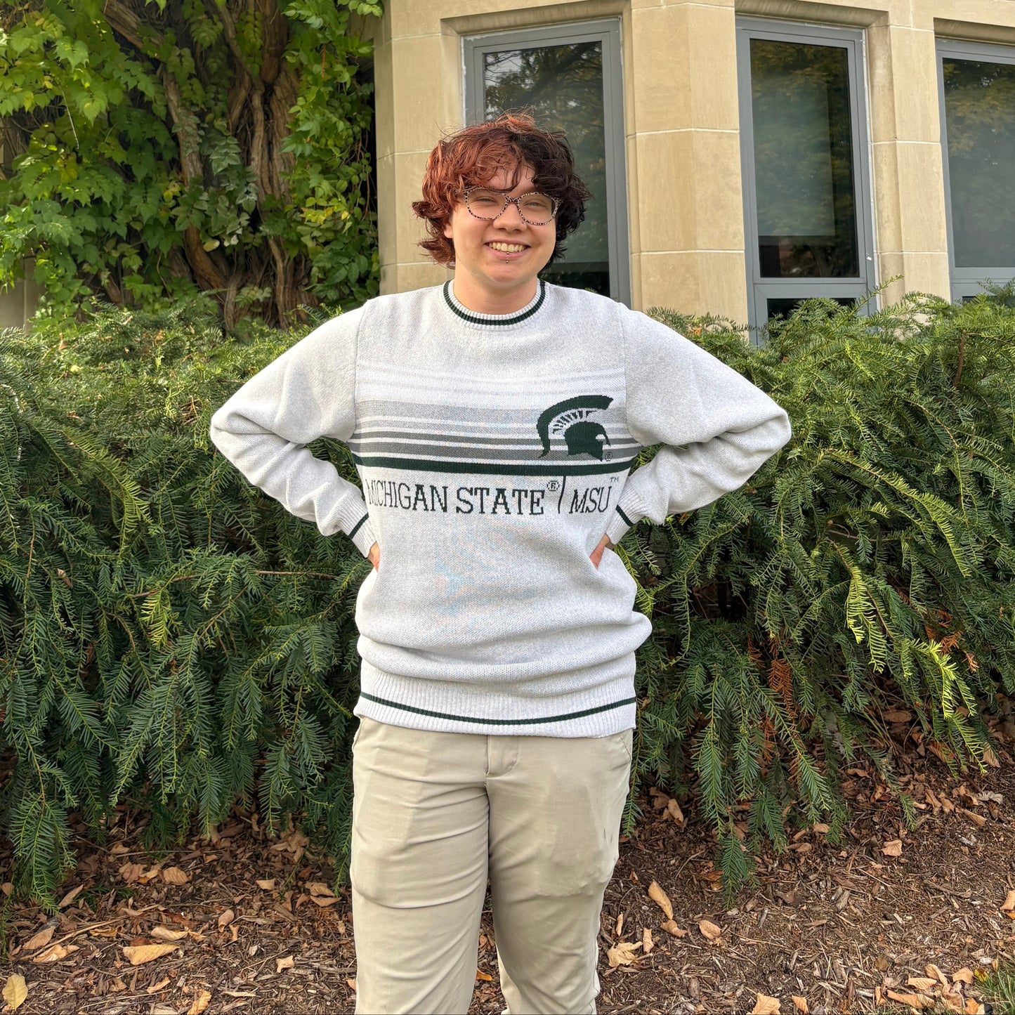 Person standing outdoors, wearing a light gray sweater with white, gray, and black stripes on the center chest section and a 
"Michigan State | MSU" wordmark in dark green below. There is a big dark green Spartan Helmet logo on the left upper chest and a dark green stripe on the collar, sleeve, and bottom hems. 