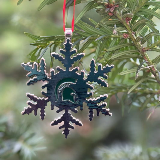 Silver snowflake-shaped metal ornament with a green Spartan Helmet logo stamped in the centered of the snowflake. Shown hanging on a tree. 