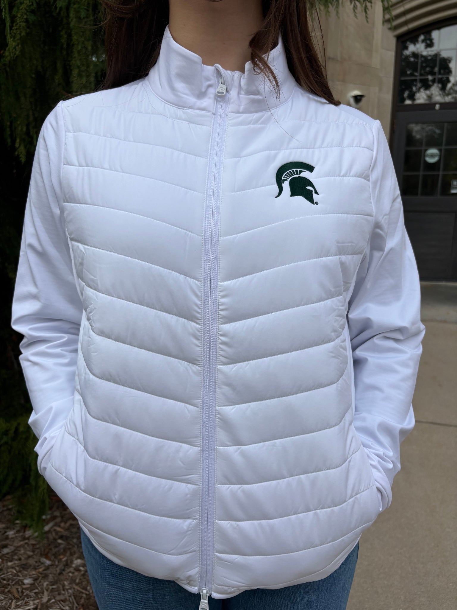 Close up of a woman standing outside, wearing a white two-way zip jacket with puff stitching on the middle section and a green Spartan helmet on the left chest. 