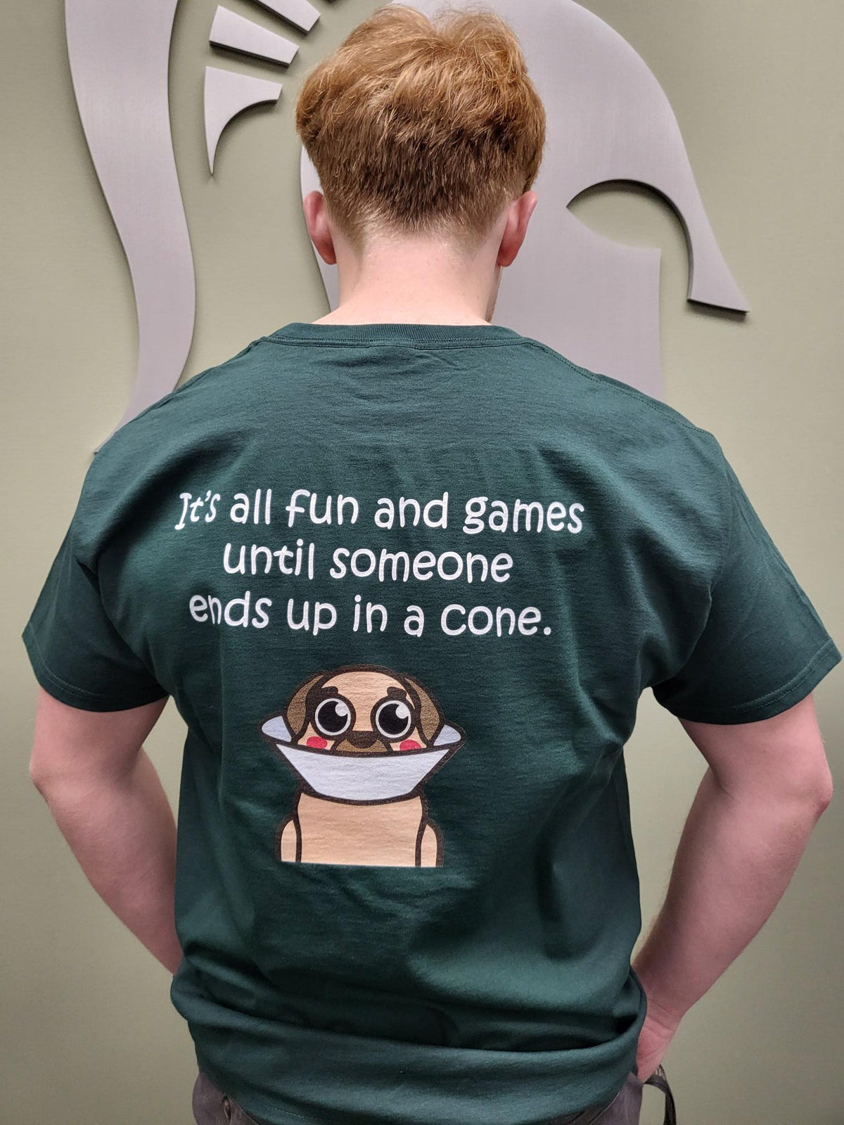 Person in a green T-shirt standing backwards. On the back of the T-shirt  in white is a wordmark that states "It's all fun and games until someone ends up in a cone." with an animated dog with a cone on below. 