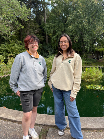 Two people standing outdoors by a body of water with trees in the background. Both people are wearing a midweight fleece button-up quarter zip. 