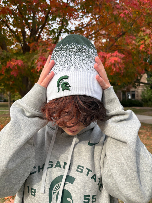Person standing outdoors, wearing a Michigan State beanie with a green and white digital gradient and the Spartan Helmet logo embroidered on the cuff in green. 