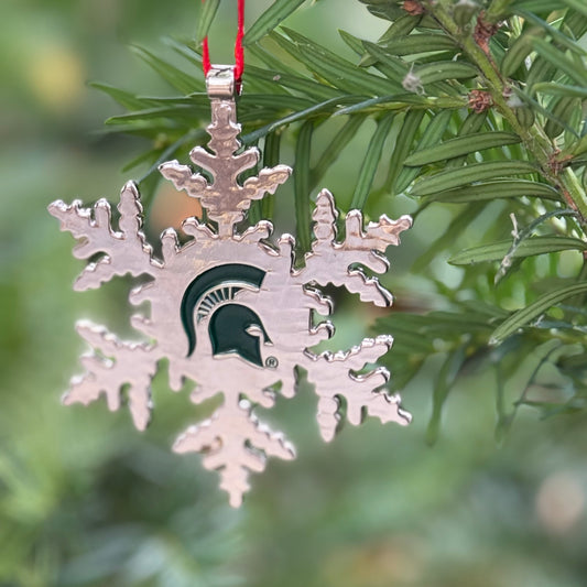 Close up of a silver snowflake-shaped metal ornament with a green Spartan Helmet logo stamped in the centered of the snowflake. Shown hanging on a tree. 