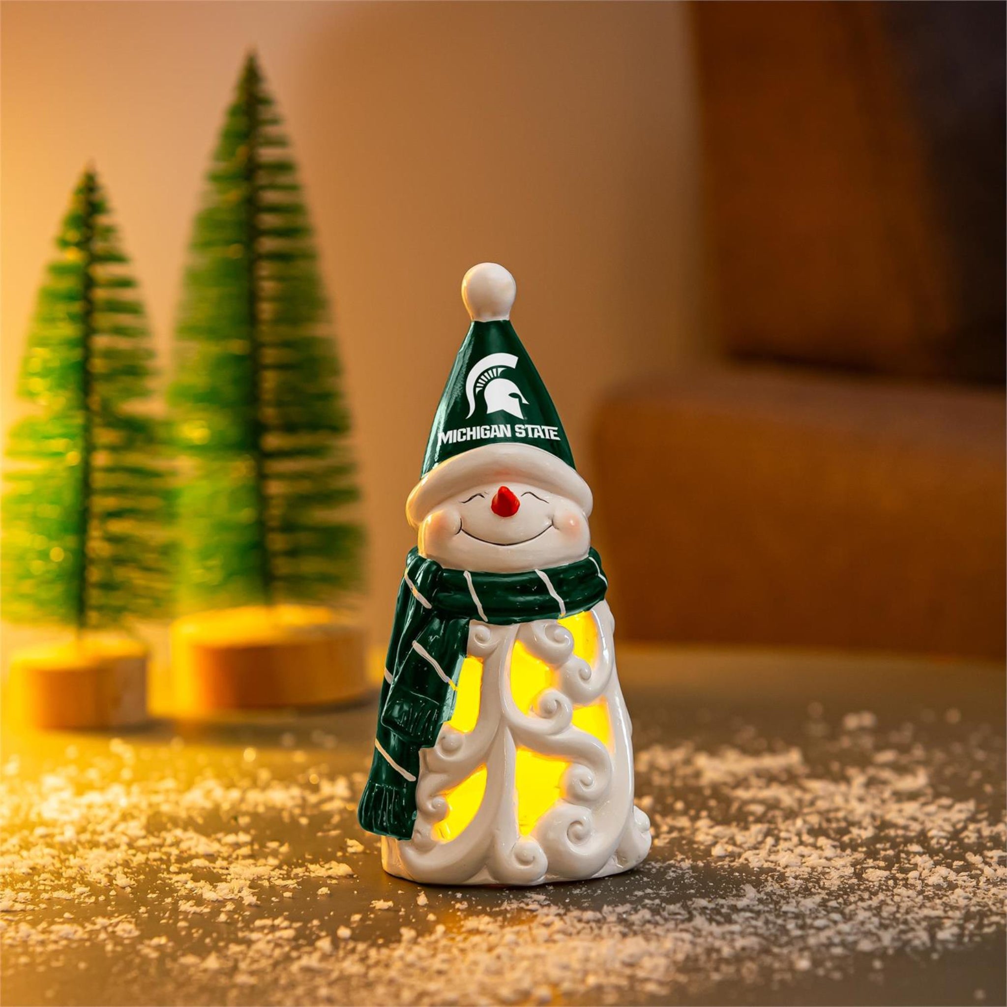 Decorative snowman figurine with a green hat that has a white Spartan Helmet in the center and white lettering that reads "MICHIGAN STATE". The snowman is also wearing a green and white striped scarf and is illuminated by a soft yellow light. Shown in a warm-toned and winter themed set.