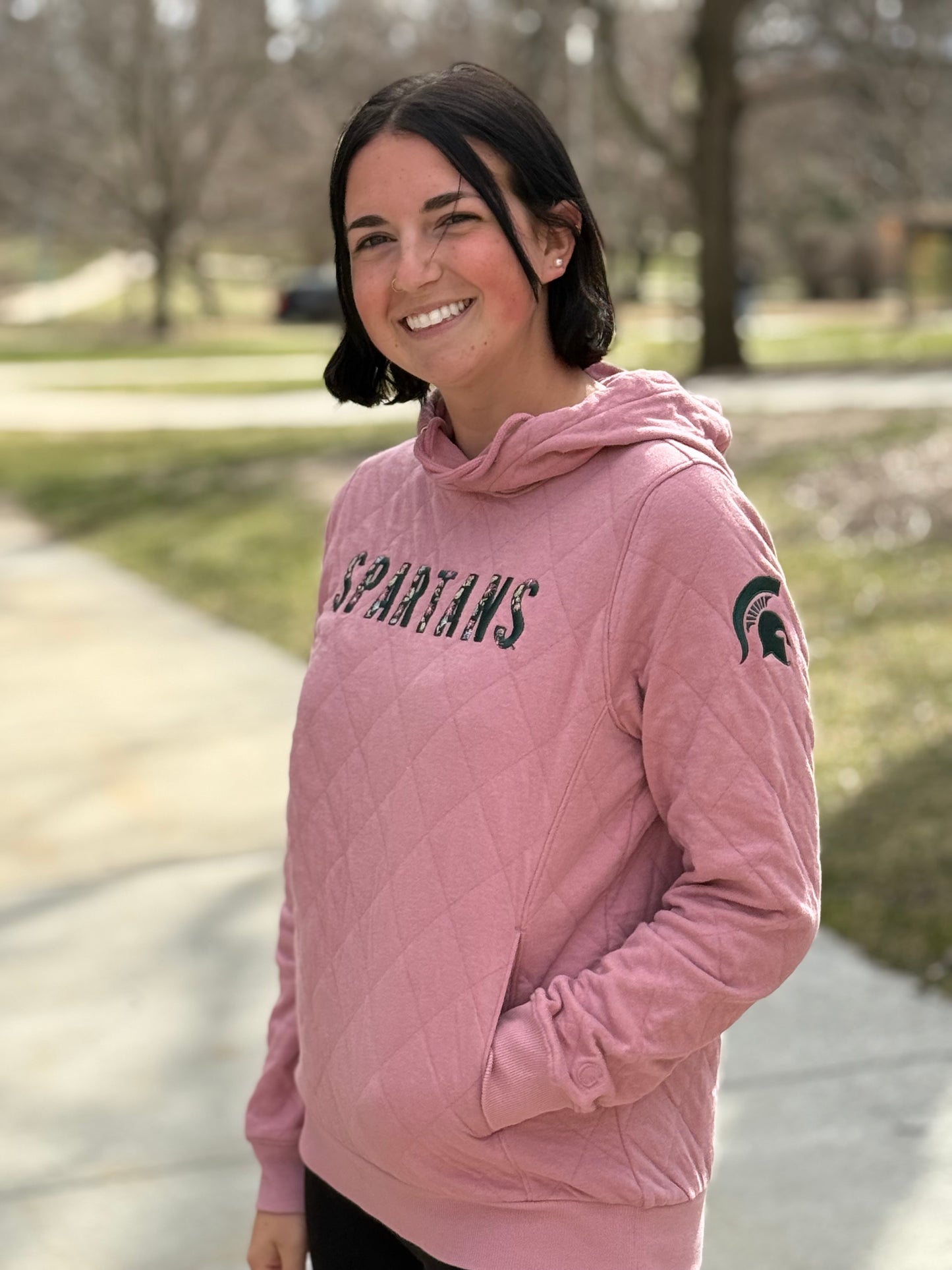 Woman standing outdoors wearing a quilted pink sweatshirt with the word "Spartans" embroidered on the front. In each letter of "Spartans" is a floral pattern. The sweatshirt has a middle pocket, drawstrings, and a green Spartan Helmet logo on the left sleeve. The sweatshirt has a middle pocket and drawstrings. 