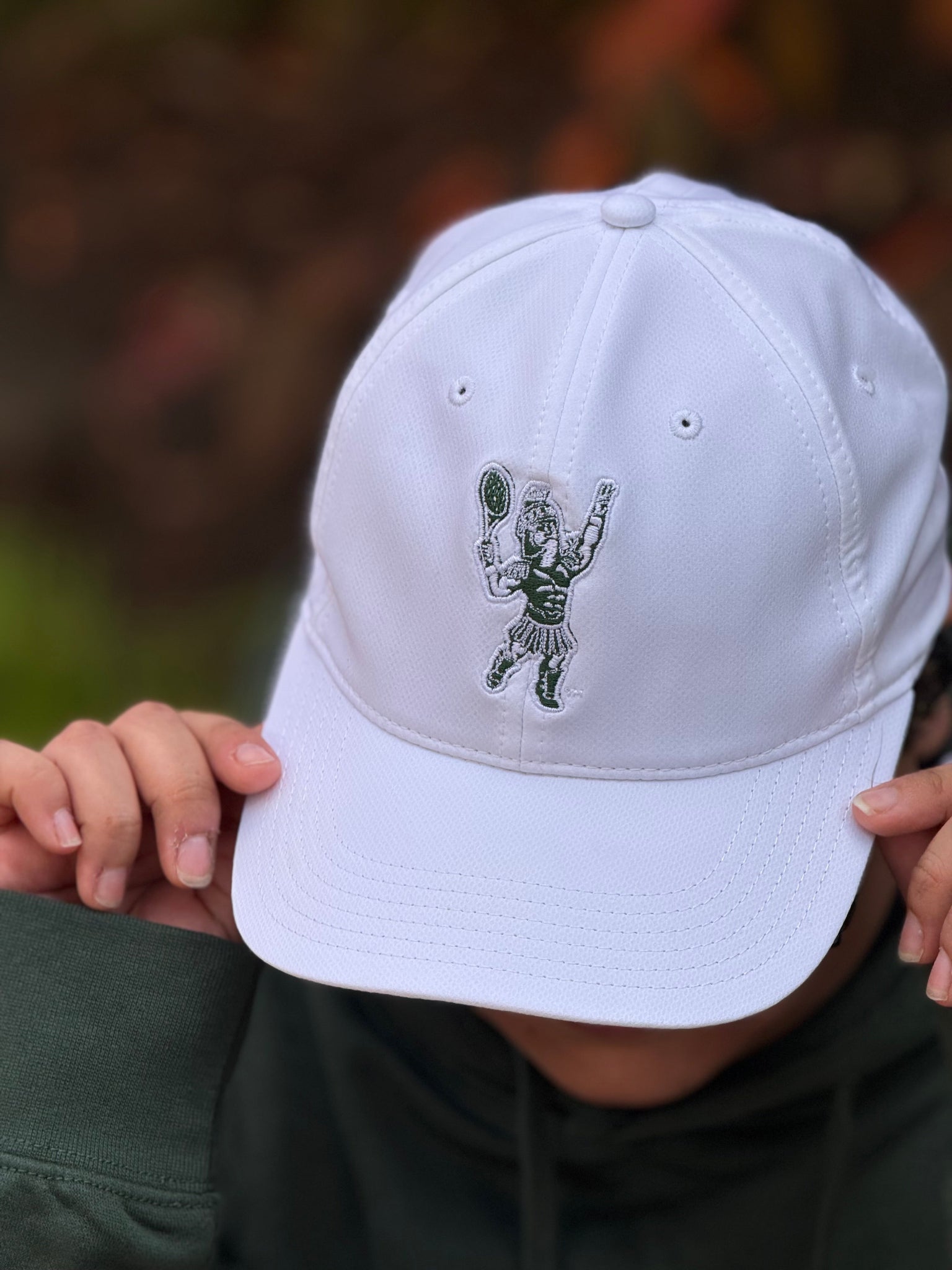 White cap with a green embroidered  Sparty playing tennis design held by a person against a blurred natural background