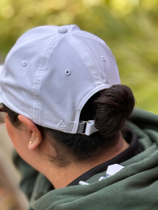 Person wearing a white cap with hair coming through the whole of the closure of hat. Shown on a blurred natural background. 