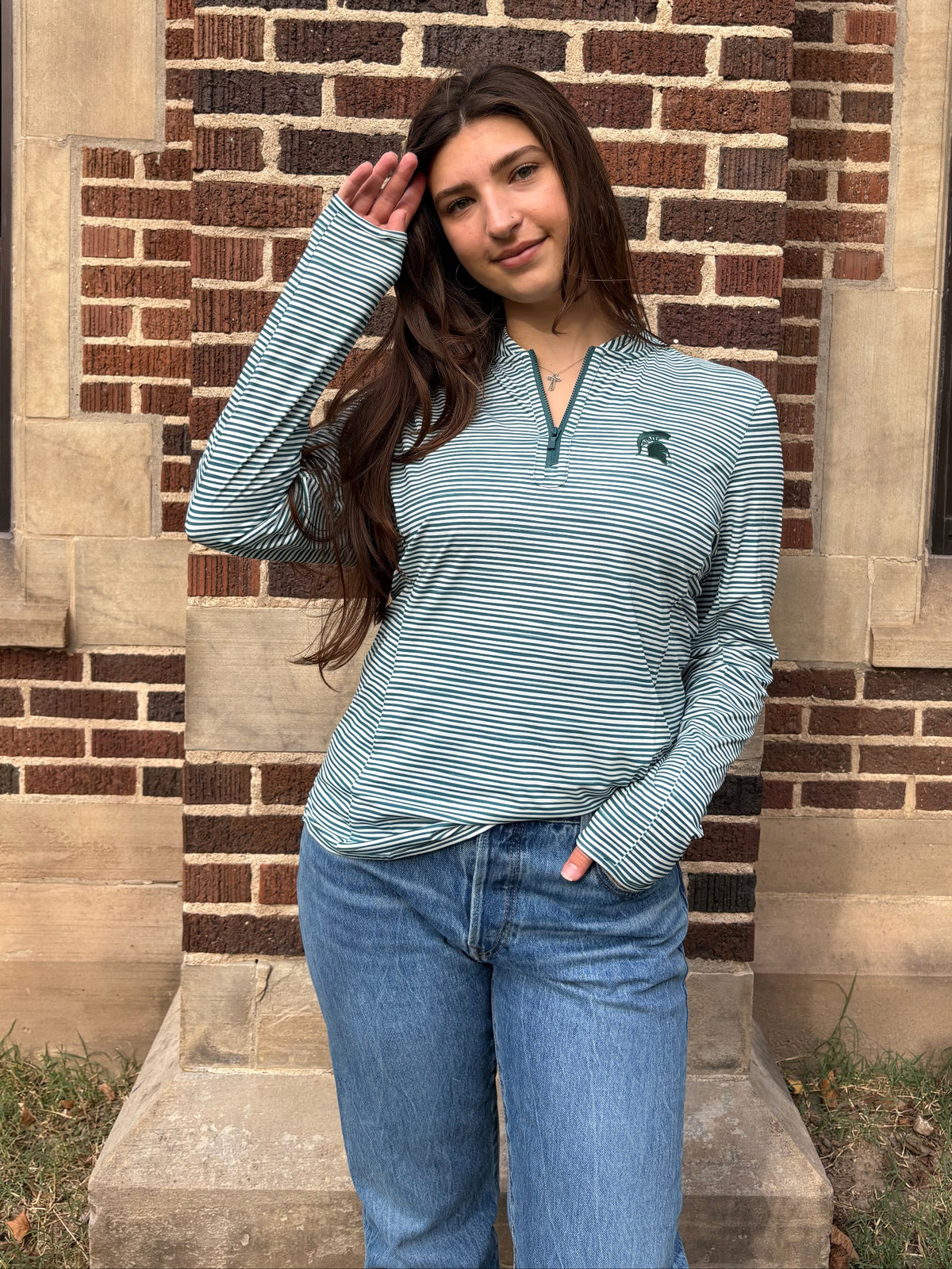 Woman standing outdoors, wearing a green and white striped quarter-zip pullover with a green Spartan Helmet logo on the left upper chest. 
