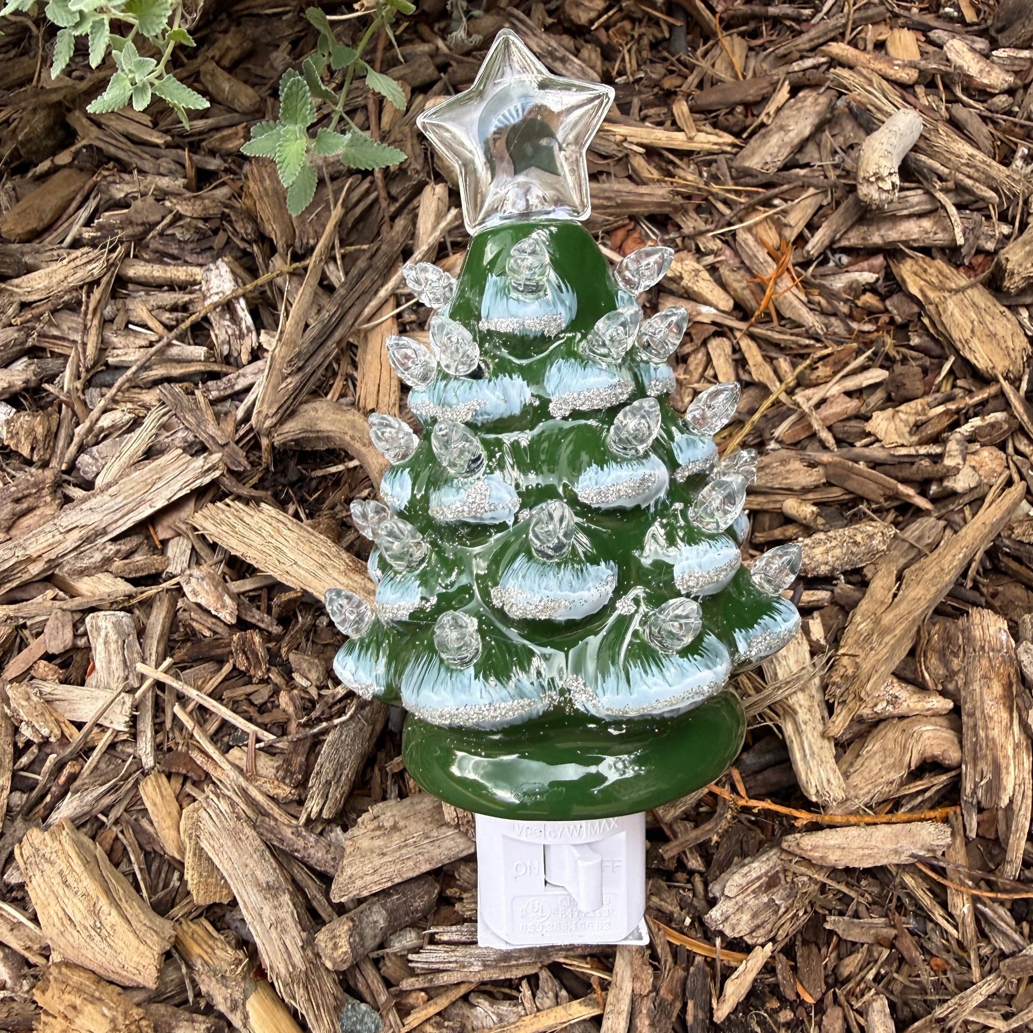 Ceramic tree night light in a evergreen tree shape with a glossy green finish, accented by painted snow tipped branches and clear bulb-style ornaments. A clear start with a green Spartan helmet logo on it is on top of the tree night light. There is a white on and off switch at the bottom of the night light. 