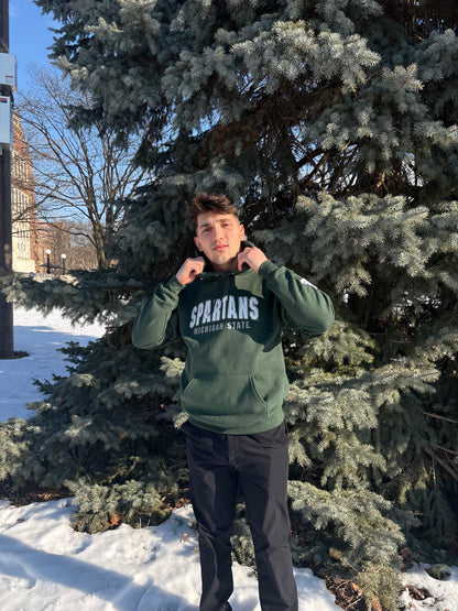 Man standing outdoors, wearing a green hoodie with "Spartans" written across the front in white and smaller white text underneath that reads "Michigan State."