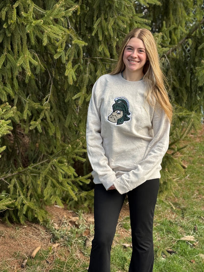 Woman wearing a oatmeal crewneck with the Gruff Sparty logo embroidered on the front chest. 