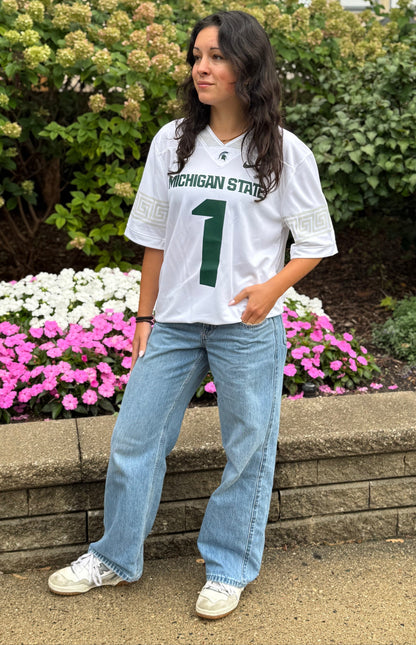 Person standing outdoors wearing a white Michigan State Spartans football-style jersey with green “MICHIGAN STATE” lettering and a large green number 1 on the front. The jersey features a Spartan helmet logo at the neckline, a Nike swoosh, and light gray geometric sleeve accents. The outfit is paired with light-wash jeans and white sneakers. Natural daylight setting with landscaped garden beds and a stone retaining wall in the background