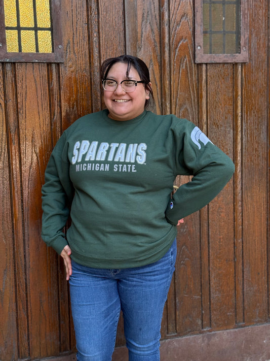Woman wearing a green crewneck with "Spartans" written in white across the front and smaller white text underneath that reads "Michigan State." On the left sleeve is a white Spartan Helmet logo. 