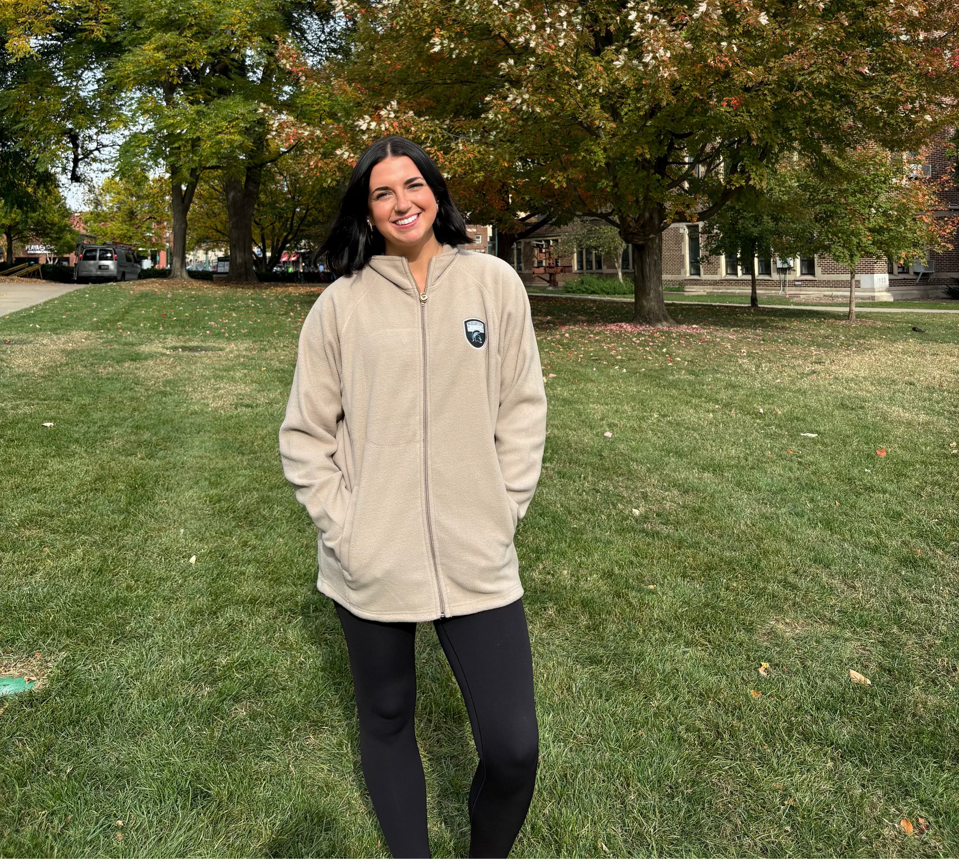 Woman standing outdoors, wearing a beige full-zip fleece jacket with a logo on the upper left chest. 
