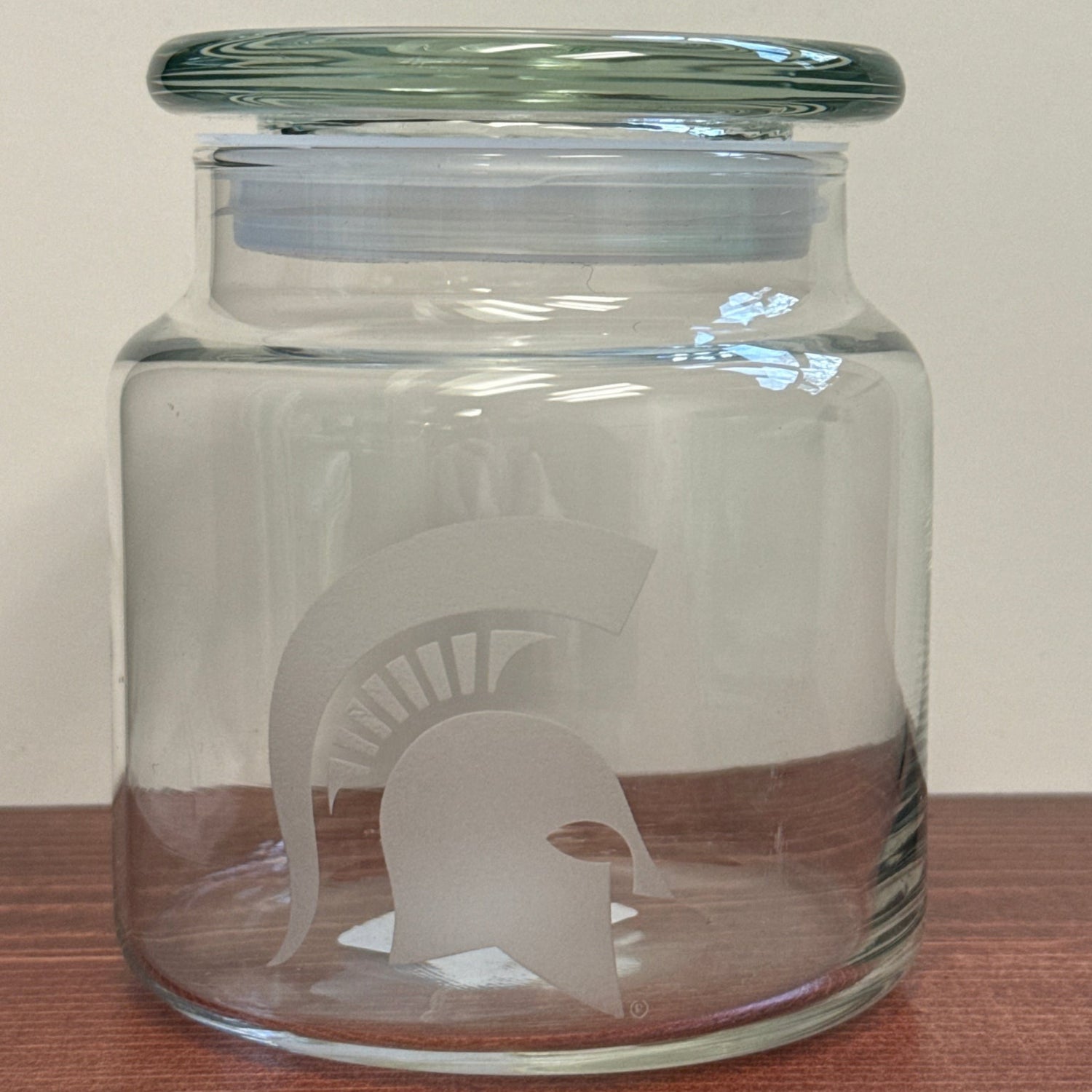 Clear glass jar with a clear lid pictured on a wooden surface and white background. Centered on the front of the jar is an acid etched white Spartan Helmet logo. 