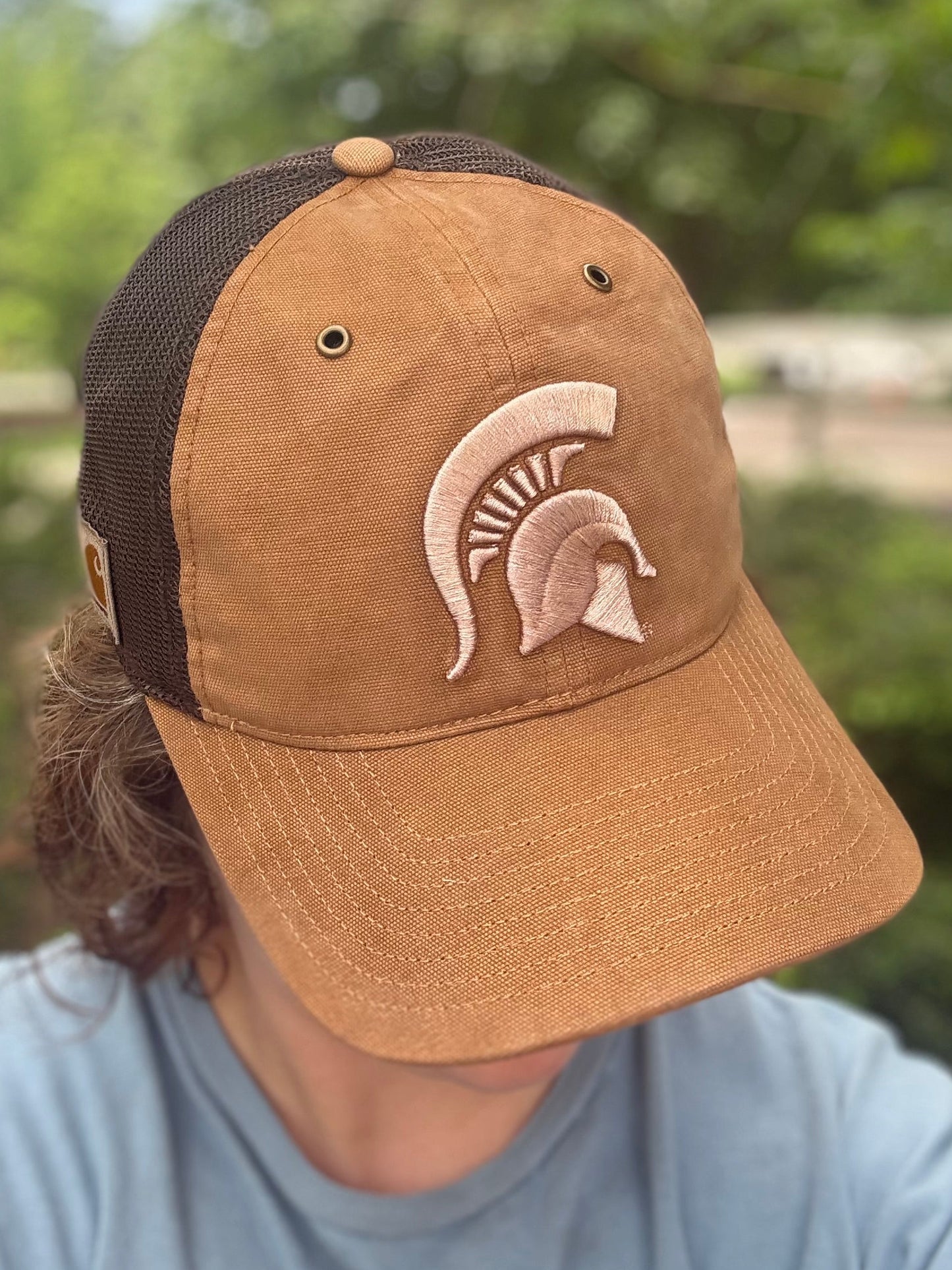 Person standing outdoors showing a brown hat with a light brown Spartan Helmet logo embroidered on the front. The sides of the hat are brown mesh. 