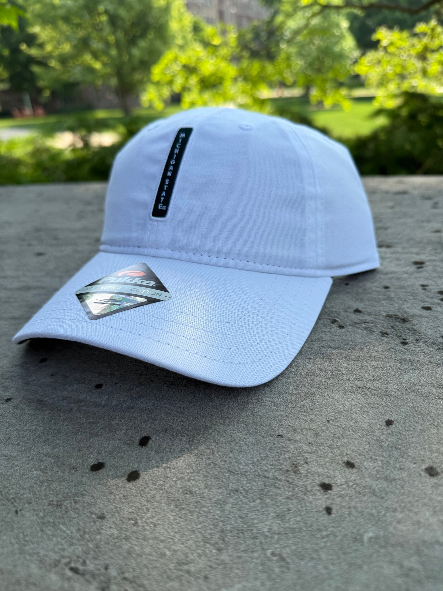 White cap with a curved bill leaning on a concrete wall outside. Green vertical bar in the center that reads Michigan State.