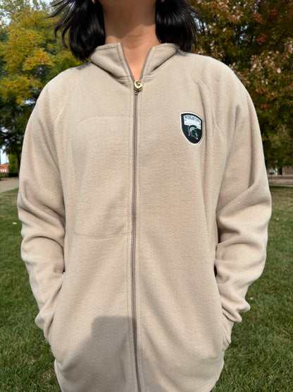 Person standing outdoors, wearing a beige full-zip fleece jacket with a logo on the upper left chest.