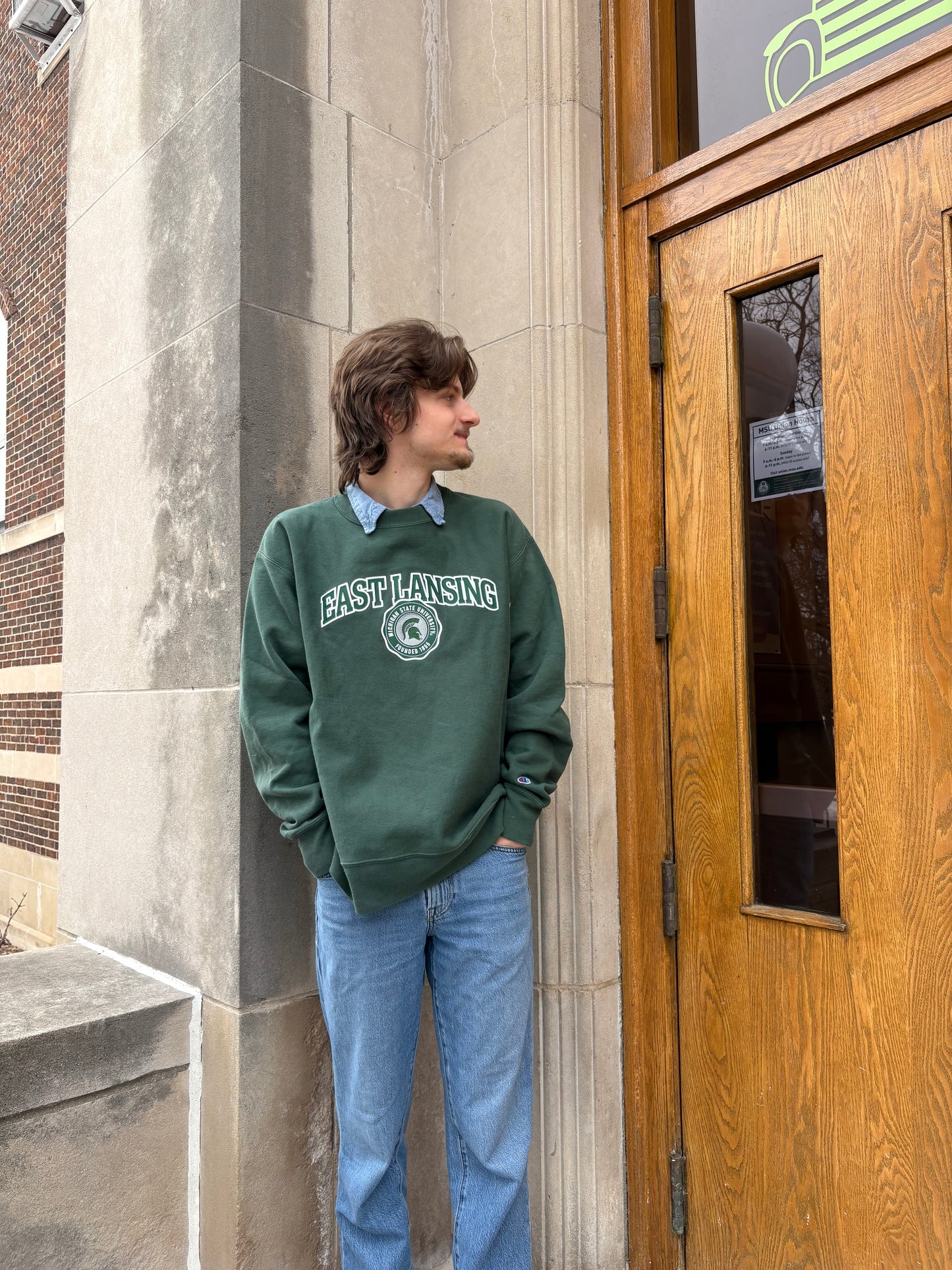 Man standing outdoors, wearing a green crewneck that has "East Lansing" written on the front chest and the Michigan State University seal logo with the Spartan Helmet logo inside, both in green and white.