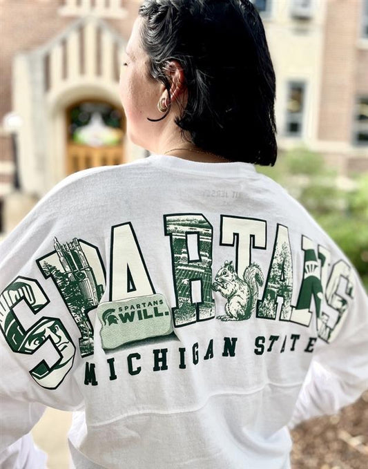 Person wearing a white Michigan State University long-sleeve shirt shown from the back, featuring oversized green-and-white “SPARTANS” lettering filled with campus-themed graphics, including landmarks, a squirrel, and the Spartan helmet logo. A green “Spartans Will.” rally towel is tucked into the back yoke, with “Michigan State” printed below the graphic.