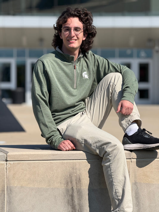 Person sitting outdoors, wearing a light green long sleeve quarter-zip with a white Spartan Helmet logo on the left upper chest and gray contrast zipper and collar.