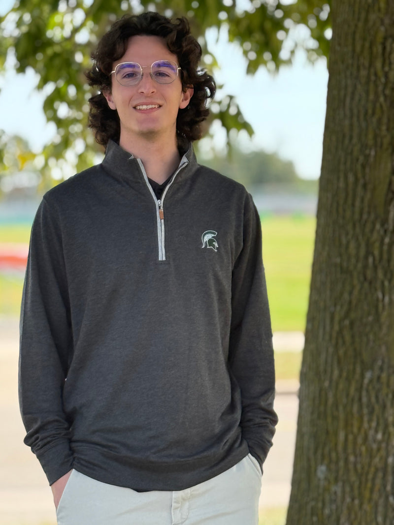Person wearing a dark gray sweatshirt with a logo, standing outdoors next to a tree.