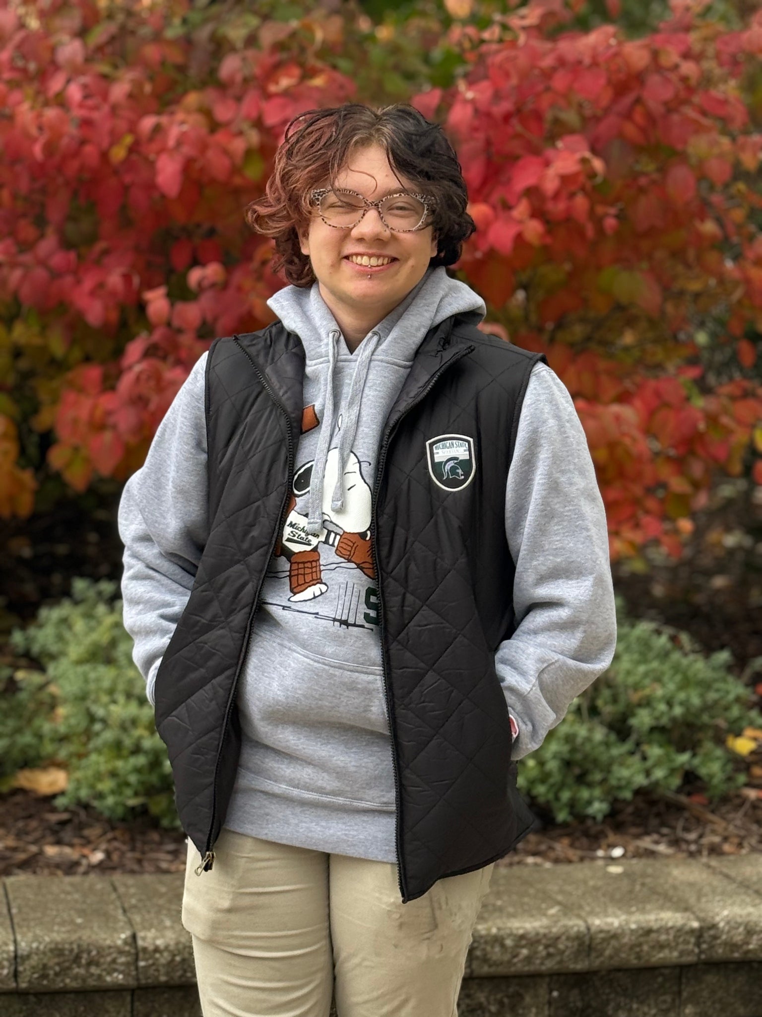 Person standing outside, wearing a full-zip quilted black vest with an official Spartans logo patch on the left upper chest. 