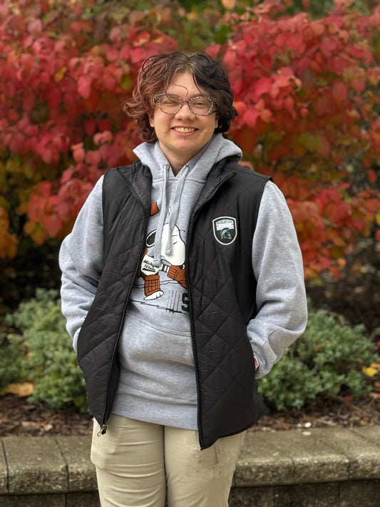Person standing outside, wearing a full-zip quilted black vest with an official Spartans logo patch on the left upper chest. 