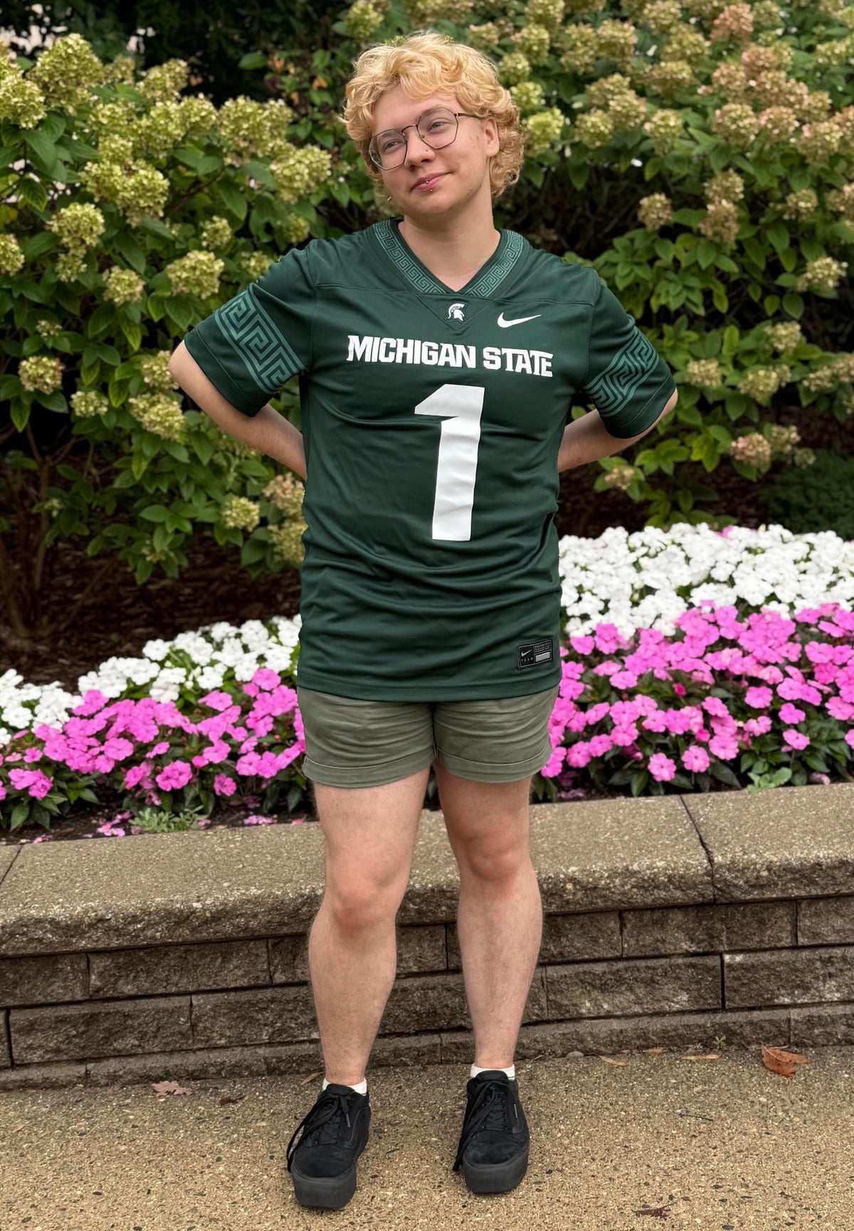 Person standing outdoors wearing a green Michigan State Spartans football-style jersey with white “MICHIGAN STATE” lettering across the chest and a large white number 1 on the front. The jersey features a Spartan helmet logo below the neckline, a white Nike swoosh, and geometric patterned sleeve accents. The outfit is paired with olive green shorts and black sneakers. A stone retaining wall and landscaped garden are visible in the background